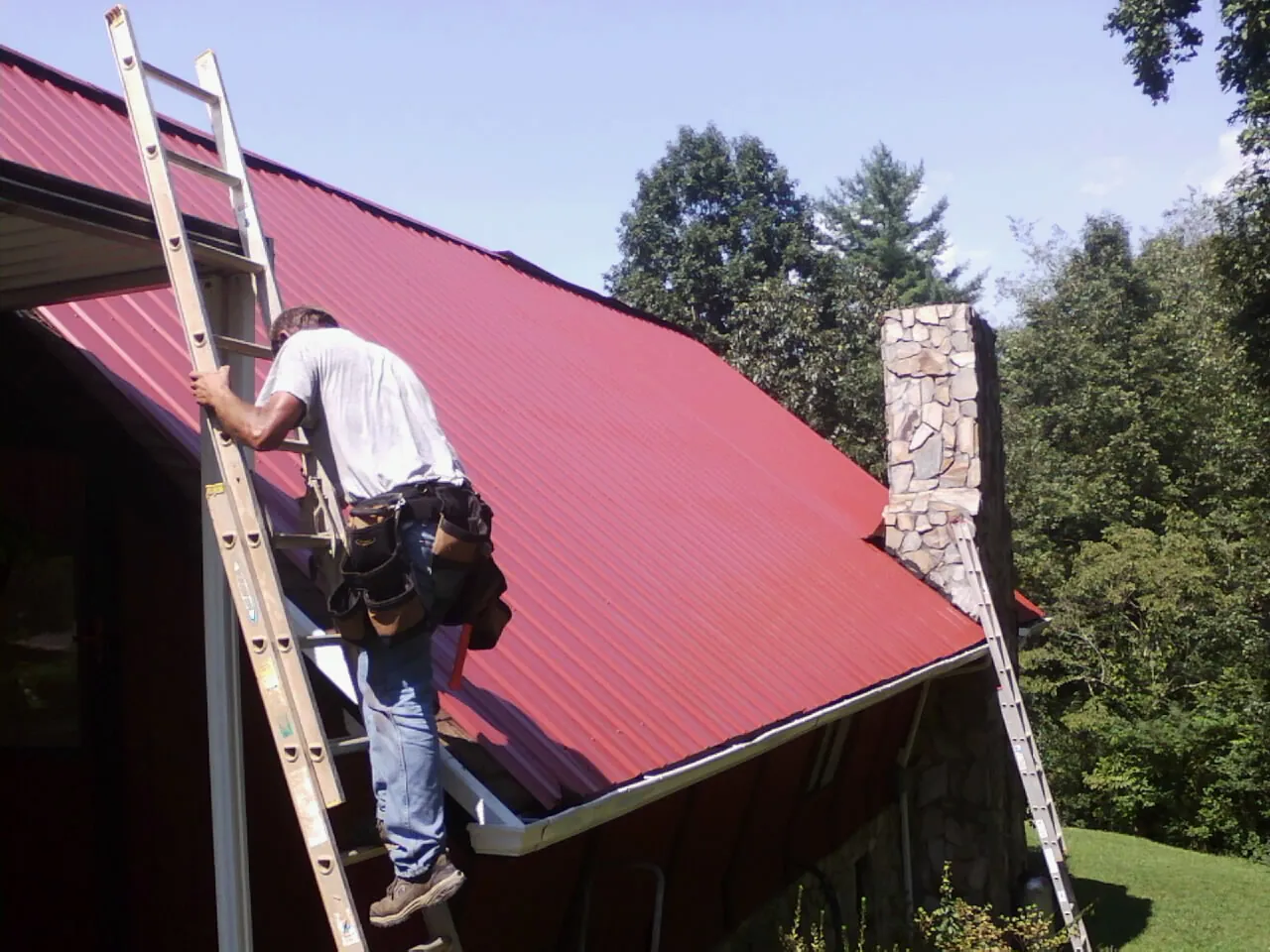 Roofer on a ladder beside a red metal roof and stone chimney — Soffit & Fascia Repair by Roofing Brothers in Waterport